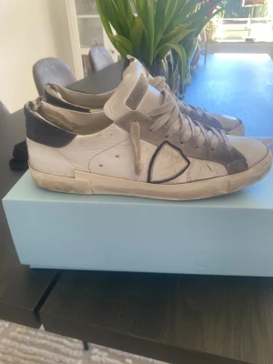 Philippe Model vita sneakers med grå detaljer - Säljer ett par Philippe Model sneakers i vitt skinn med grå mockadetaljer och svarta inslag. Klassisk låg modell med snörning och gummisula. Ikonisk sköld på sidan och Philippe Model-logga på sulan. Skorna levereras med originalkartong och dustbag.