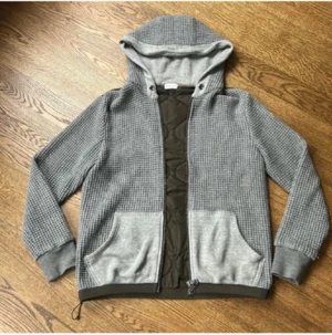 Grå quiltad hoodiejacka från Moncler - Snygg grå hoodiejacka från Moncler med quiltad mörkbrun panel på ryggen och insidan. Jackan har huva, två stora fickor fram och dragkedja. Stickad struktur på ärmar och huva, samt ribbade muddar. Perfekt för dig som gillar streetwear med exklusiv känsla.