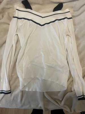 Offshoulder vit stickad tröja - Snygg offshoulder stickad tröja i vit med svarta ränder längs halsringningen och ärmsluten. Tröjan har lång ärm och ribbade muddar, perfekt för dig som gillar en stilren look med en twist. Passar dig som vill ha något lite extra till garderoben.