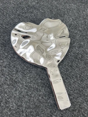 Silvrig hjärtformad sminkspegel - Säljer en silvrig sminkspegel i hjärtform med coolt vågig yta. Spegeln har ett handtag som gör den enkel att använda när du fixar sminket. Perfekt för att få till detaljerna och ta snygga selfies.