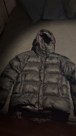 Svart pufferjacka från Haglöfs - Svart pufferjacka från Haglöfs med huva och markerade quiltade sektioner. Jackan har dragkedja framtill och är tillverkad i syntetmaterial. Perfekt för kalla dagar och har en clean, sportig look.