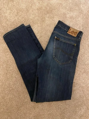 DarkBlue Lee Jeans - Mörkblåa Jeans från Lee  Klassisk och stilrena jeans i en neutral svart nyans. Modellen har ett Lee märke vid högra bakfickan, vilket ger en diskret look. Perfekt både till vardags och lite finare tillfällen.  🔹 Storlek: se bild 2/3 🔹 Färg: Mörkblå 🔹 Skick: Mycket fint skick – inga synliga defekter, hål eller fläckar. 🔹 Material: Kontrollera gärna tvättlappen (ej synlig på bilden). 🔹 Nypris: Ca 1 400 - 1 700kr beroende på modell och butik  Ett par tidlösa byxor som passar dig som gillar klassisk