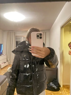 Calvin klein jacka - Säljer en svart, glansig pufferjacka från Calvin Klein Jeans med stor huva och dragkedja framtill. Jackan har quiltad design och logga på bröstet. Materialet är syntet och polyester.