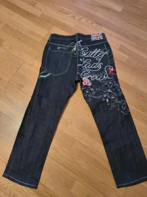 Artful Dodger jeans med broderi - Unika mörkblå jeans från Artful Dodger med broderade detaljer, färgglada sömmar och coolt motiv på bakfickan. Brittisk flagga på linningen och broderad text samt blommor och figurer ger en riktigt personlig vibe. Passar dig som vill sticka ut.