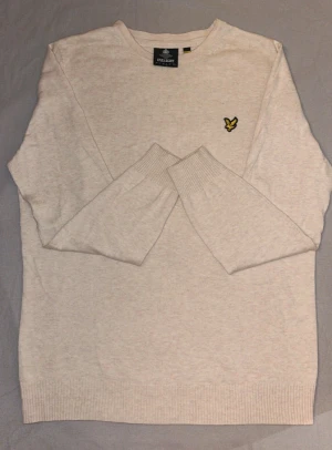 Beige stickad tröja från Lyle & Scott - Snygg beige stickad tröja från Lyle & Scott med klassisk rund hals och deras ikoniska gula logga broderad på bröstet. Tröjan har långa ärmar och ribbade muddar för en clean look. Perfekt för lager-på-lager under kyliga dagar.