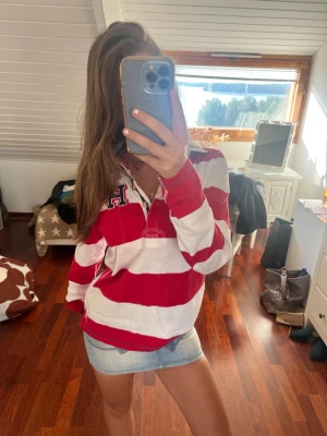 Rödvit randig Henley från Tommy Hilfiger - Säljer en snygg Henley-skjorta från Tommy Hilfiger i storlek L. Skjortan har breda röda och vita ränder, klassisk krage och knappar vid halsen. På bröstet finns ett broderat H och emblem. Materialet är mjuk bomull, perfekt för en chill och klassik look.