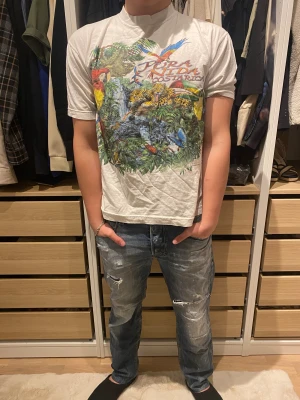 Vit T-shirt från Costa rica - Säljer en vit t-shirt från Tico Soul med färgglatt Costa Rica-tryck på framsidan. Motivet har djungel o olika exotiska djurr. T-shirten är kortärmad och har en klassisk passform. Materialet är mjuk bomull, perfekt för dig som gillar unika prints.