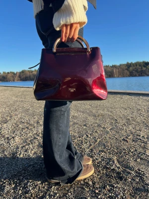 Vinröd handväska från Dune London - Snygg vinröd handväska från Dune London med glansig finish och stilrena metallhandtag i guld. Väskan har en elegant, boxig form och är perfekt för dig som vill ha en chic accessoar med lyxig vibe. Färgen är djup vinröd med metalliska detaljer.