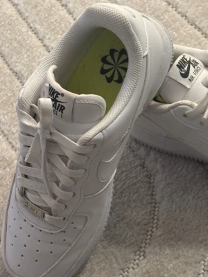 Nike Air Force 1 vita sneakers - Klassiska Nike Air Force 1 sneakers i helvitt skinn med perforerad tå, snörning och ikonisk sula. Innersulan är limegrön med svart blomlogga. Perfekta för dig som gillar en clean och tidlös look.