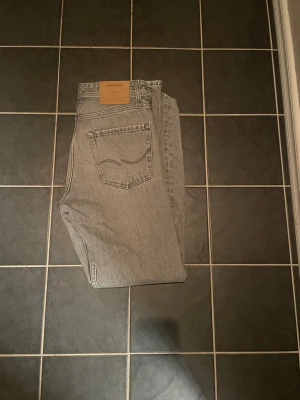 Grå jeans från Jack & Jones - Snygga grå jeans från Jack & Jones med klassisk femficksdesign och raka ben. Jeansen har en ljusgrå tvätt och är tillverkade i slitstarkt bomullsjeansmaterial. Passar dig som gillar en enkel och stilren look med diskreta detaljer på bakfickorna.