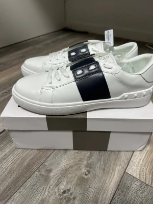Valentino Garavani sneakers vit/blå - Snygga Valentino Garavani sneakers i vitt skinn med bred mörkblå rem över vristen och coola nitar på hälen. Klassisk rund tå, platt sula och vita snören. Märkeslogga i guld på plösen. Perfekt för dig som gillar exklusiv streetstyle.