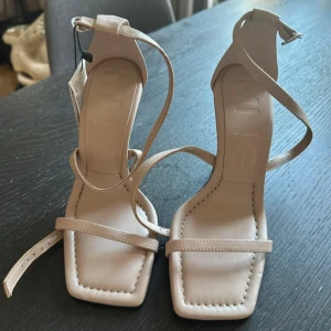 Beige sandaler med smala remmar och klack - Stilrena beige sandaler från Mango med smala remmar, fyrkantig tå och låg klack. Skorna har justerbart spänne runt vristen. Perfekta för dig som gillar minimalistisk och trendig design.