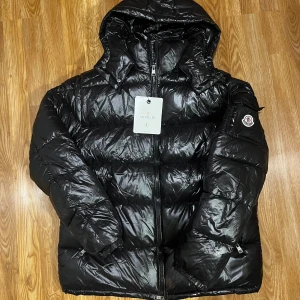 Svart pufferjacka från Moncler - Säljer en svart pufferjacka från Moncler med huva och klassisk logga på ärmen. Jackan har glansig yta, dragkedja framtill och quiltad design som ger en cool look. Perfekt för kalla dagar och riktigt snygg till streetwear.