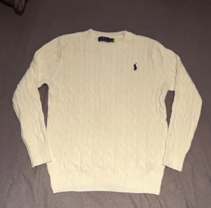Vit kabelstickad tröja Polo Ralph Lauren - Säljer en klassisk vit kabelstickad tröja från Polo Ralph Lauren i storlek S. Tröjan har rund hals, lång ärm och den ikoniska broderade loggan på bröstet. Tillverkad i 100% bomull för en mjuk och skön känsla. Perfekt för dig som gillar stilrena och tidlösa plagg.