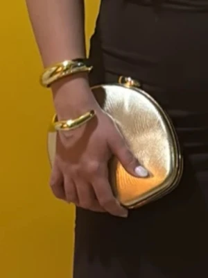 Guldig kuvertväska  - Snygg kuvertväska i guldigt syntetmaterial med vågigt mönster och rundad form. Väskan har ett elegant snäpplås upptill och en avtagbar guldfärgad kedja.