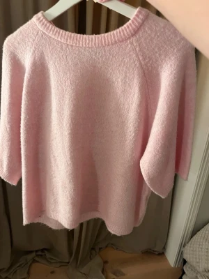 Ljusrosa stickad tröja från 157 - Mysig ljusrosa stickad tröja från 157 med rund hals och ribbad kant. Tröjan har en loose passform och trekvartsärm, perfekt för dig som gillar en chill och söt stil. Materialet är mjukt och fluffigt, vilket gör den extra skön att bära.