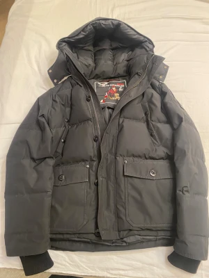 Svart Moose Knuckles dunjacka - Svart dunjacka från Moose Knuckles med stor huva, ribbade muddar och flera fickor med knappar. Jackan har dragkedja och knappar framtill, samt ikonisk logga på ärmen. Perfekt för kalla dagar och har en schysst, lite loose passform. Pris kan diskuteras