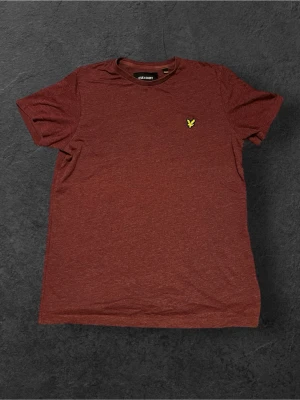 Röd t-shirt från Lyle & Scott - Snygg röd t-shirt från Lyle & Scott med klassisk gul fågel-logga broderad på bröstet. T-shirten har rund hals och korta ärmar, perfekt för en chill och stilren look. Materialet är mjuk bomull som känns skönt mot huden.