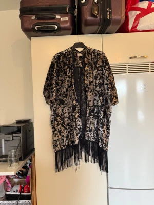 Svart och silver kofta med fransar Cream - Unik kofta från Cream i storlek M/L. Men den är mer en L/XL., Svart och silvermönster och snygga fransar längs nederkanten. Oversized passform och korta ärmar, perfekt för att ge din outfit en cool touch. Materialet är mjukt och har en glansig finish. Mycket fint skick, jag hittar inga synliga anmärkningar eller defekter. 