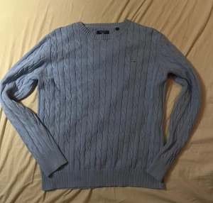 Ljusblå kabelstickad tröja från GANT - Säljer en ljusblå kabelstickad tröja från GANT i storlek L. Tröjan är tillverkad i mjuk bomull och har ett klassiskt kabelmönster samt rund halsringning. GANT-loggan med broderad sköld finns på bröstet. Perfekt för dig som gillar preppy stil.