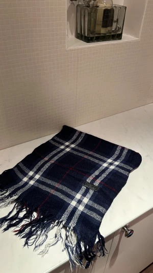 Blå rutig ullväst från Burberry - Snygg blå väst från Burberry med klassiskt rutigt mönster i vitt och rött. Västen är gjord av 100% lammull och har fransar längs nederkanten. Perfekt för dig som gillar tidlös stil och vill ha något som sticker ut lite extra.