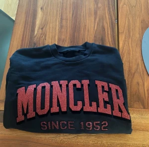 Svart Moncler sweatshirt med röd text - Svart sweatshirt från Moncler med stor röd  text 'MONCLER SINCE 1952' på bröstet. Tröjan har rund hals, långa ärmar och ribbade muddar. Tillverkad i mjuk bomull för skön komfort och stilren look.
