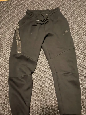 Svarta Nike teach set med snörning passar S - Svarta mjukisbyxor från Nike i storlek M med snörning i midjan och dragkedjedetalj på ena benet. Byxorna har en stilren look med Nike-logga på låret och är gjorda i mjukt bomullsmaterial. Perfekt för chill eller träning. Passar även storlek (S)