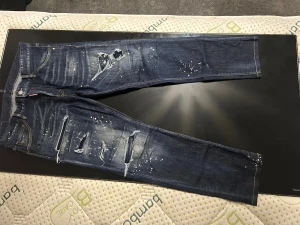 Mörkblå Dsquared2 jeans med slitningar - Säljer ett par mörkblå jeans från Dsquared2 med coola slitningar, färgstänk och distressed detaljer både fram och bak. Klassisk femficksmodell med gul patch bak och kontrastsömmar. Perfekt för dig som gillar streetwear och vill sticka ut.