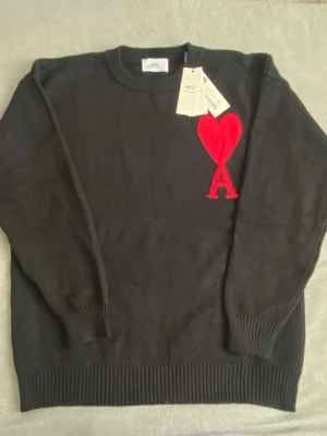 Svart stickad tröja från Ami med hjärta - Svart stickad tröja från Ami Alexandre Mattiussi med rund hals och ribbade muddar. På bröstet finns ett stort rött hjärta och bokstaven A som broderad detalj. Perfekt för dig som gillar stilrena plagg med en twist. Passar till både M och L 
