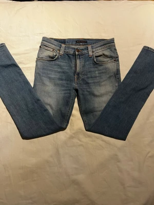 Blå jeans från Nudie Jeans - Snygga blå jeans från Nudie Jeans Co i klassisk femficksmodell. Jeansen har en rak passform, orangea sömmar och Nudie-logga på bakfickan. Materialet är slitstarkt denim och färgen är ljusblå med lätt tvättad look. Perfekt för dig som gillar stilrena och tidlösa jeans. Modellen är Thin Finn i storlek 32/32.
