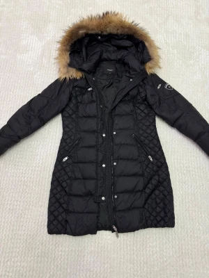 Svart lång dunjacka Rockandblue - Snygg svart lång dunjacka från Rockandblue med quiltade partier, pälskantad huva och flera dragkedjor. Jackan har en figurnära passform, emblem på ärmen och praktiska fickor. Perfekt för kalla dagar när du vill vara både varm och stilren.