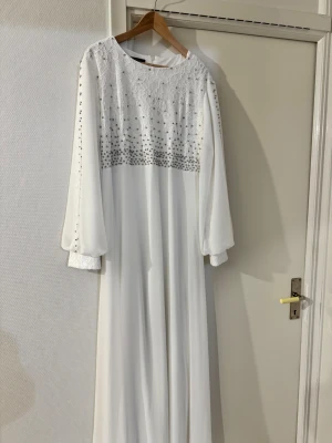Vit långklänning med spets och pärlor - Elegant vit långklänning med spets upptill och pärldetaljer över byst och ärmar. Klänningen har långa, lösa ärmar med spetsmuddar och pärlor längs hela ärmen. Kjolen är i ett lätt, flödande tyg som ger en drömmig känsla. Har bara använts 1 gång i 4h.