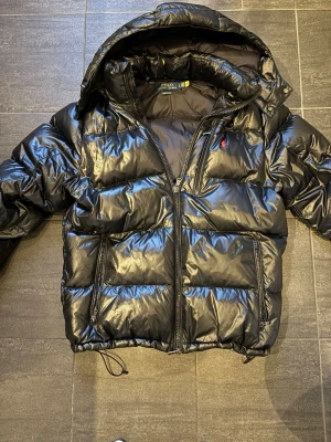 Svart pufferjacka från Polo Ralph Lauren - Svart pufferjacka från Polo Ralph Lauren med huva och dragkedja framtill. Jackan har två sidofickor med dragkedja och den klassiska röda Polo-loggan på bröstet. Materialet är glansigt och jackan har justerbar dragsko nedtill för extra komfort. Jackan är nästintill oanvänd och har inga defekter. PRIS KAN DIKSUTERAS
