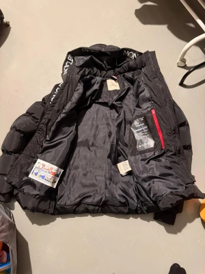Svart Moncler dunjacka med huva - Säljer en svart Moncler dunjacka med huva och logotyp på insidan av kragen. Jackan är vadderad med gåsdun, har dragkedja framtill och praktiska fickor. Snygg och klassisk modell som håller dig varm under kalla dagar. Har använt några gånger 