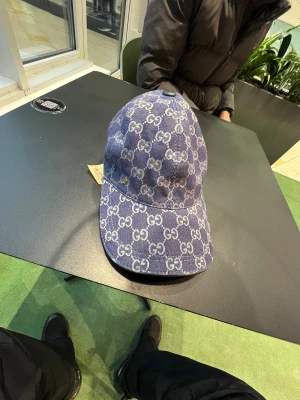 Blå Gucci keps i denim med GG-mönster - Säljer en blå keps från Gucci i denim med det klassiska GG-monogrammet över hela kepsen. Justerbar rem baktill i skinn och insida i ljus bomull. Snygg streetstil och perfekt för dig som gillar exklusiva accessoarer. Äkta! 