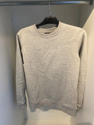 Nike Sweatshirt  - Snygg grå sweatshirt från Nike i storlek S. Tröjan har rund hals, långa ärmar och ett litet vitt Nike-logotryck på bröstet. Tillverkad i mjuk bomullsmix med ribbade muddar vid ärmslut och nederkant. Perfekt för chill eller träning.                                              Obs den är en (Rep)
