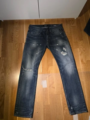 Replay Newbill jeans blå, storlek 32 - Säljer ett par Replay Newbill jeans i blå tvätt med slitna detaljer och snygga rippade partier. Klassisk femficksmodell med raka ben och normal passform. Jeansen har dragkedja och knapp i midjan, samt Replay-logga på insidan.