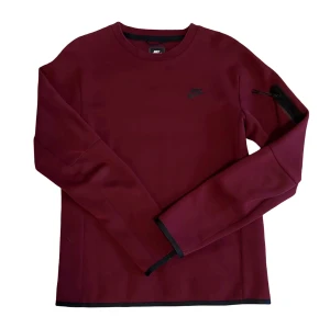 Nike Tech Fleece sweatshirt  - Snygg vinröd långärmad tröja från Nike med svart logga på bröstet och en diskret ficka med dragkedja på ärmen. Tröjan har rund hals och svarta detaljer vid muddar och nederkant. Materialet är mjukt med 66% bomull och 34% polyester.