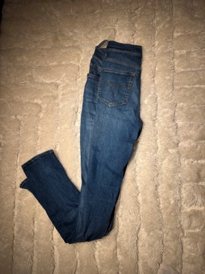 Blå jeans 29/32 från T/J - Snygga blå jeans från T/J i modellen Kelly, storlek 29/32. Klassisk femficksdesign med bruna sömmar och läderpatch bak i midjan. Jeansen har rak passform och är tillverkade i slitstarkt denimtyg. Perfekta för dig som gillar en enkel och tidlös look. En skärpföstare har släppt, kan tas bort eller nog sy tillbaka 