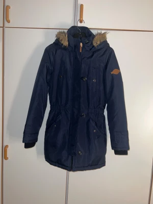 Marinblå parkas från Vero Moda - Snygg marinblå parkas från Vero Moda med huva och fuskpäls. Jackan har dragkedja och knappar framtill, två stora fickor och ribbade muddar vid ärmslut. Perfekt för kalla dagar med sin långa passform och justerbar midja.
