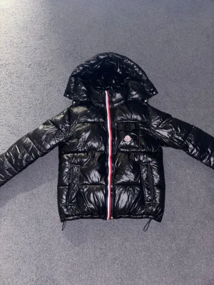 Svart Moncler pufferjacka med huva - Säljer en svart glansig pufferjacka från Moncler med huva och klassisk logga på bröstet. Jackan har dragkedja med röd, vit och blå detalj, flera fickor och quiltad design. Perfekt för kalla dagar när du vill ha både stil och värme.