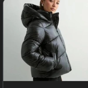 Svart pufferjacka med huva - Säljer en svart, glansig pufferjacka med huva och dragkedja framtill. Jackan har en oversized passform, elastiska muddar vid ärmslut och stora fickor på sidorna. Perfekt för kalla dagar när du vill hålla värmen och ändå se cool ut.
