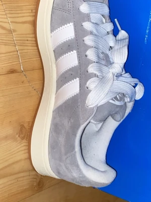 Adidas Campus grå sneakers mocka - Adidas Campus sneakers i ljusgrå mocka med klassiska vita ränder på sidorna och vit text. Skorna har vit platt sula och gummisula i brun färg. Tjocka vita snören och rund tå ger en clean och stilren vibe. Perfekt för dig som gillar retro och streetwear.
