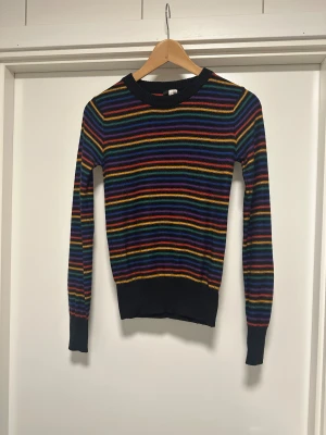 Randig stickad tröja i regnbågsfärger - Snygg stickad tröja med färgglada ränder i lila, blått, grönt, gult, orange och rött mot svart bakgrund. Tröjan har rund hals, ribbade muddar och lång ärm. Perfekt för dig som gillar färg och vill sticka ut lite extra. 