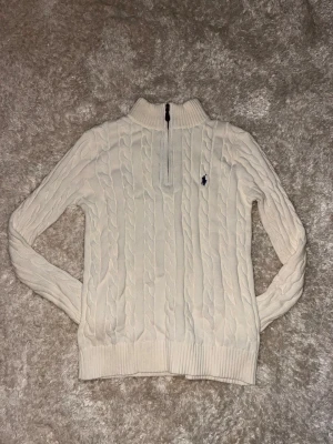 Vit stickad half zip tröja Polo Ralph Lauren - Säljer en vit stickad tröja från Polo Ralph Lauren med half zip och kabelstickat mönster. Tröjan har långärmad design, ribbad krage och muddar samt den klassiska blå Polo-loggan broderad på bröstet. Perfekt för dig som gillar clean och stilren look.
