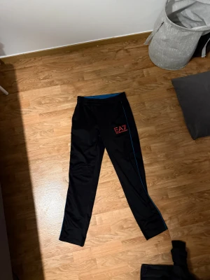 Svarta EA7 Emporio Armani byxor - Säljer ett par svarta träningsbyxor från EA7 Emporio Armani med röd logga på vänster lår och blå detaljer längs benet. Byxorna har elastisk midja och är tillverkade i ett syntetmaterial som är skönt och lätt. Perfekt för dig som gillar sportig stil.