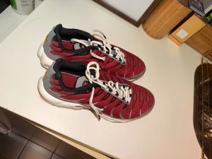 Nike Air Max Plus Tn röda sneakers - Nike Air Max Plus Tn sneakers i rött och grått med svarta detaljer och vit sula. Skorna har klassisk snörning, mesh-överdel och Tn-logga bak. Snyggt vågigt mönster på sidorna och bekväm platt sula med synlig Air Max-dämpning.