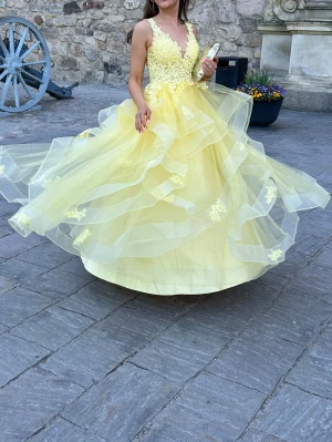 Gul långklänning med spets och volanger - Säljer en gul balklänning från Jj’House med v-ringad urringning, spetsdetaljer på överdelen och flera lager volanger i kjolen. Klänningen har ärmlös design och är tillverkad i tunn tyll med broderade blommor.