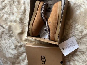 UGG bruna mocka tofflor  - Säljer ett par bruna slip-on skor från UGG i mjuk mocka med fluffigt foder och grov sula. Skorna har ett snyggt broderat mönster runt öppningen i vitt och svart. Perfekta för chill dagar och håller fötterna varma och sköna.