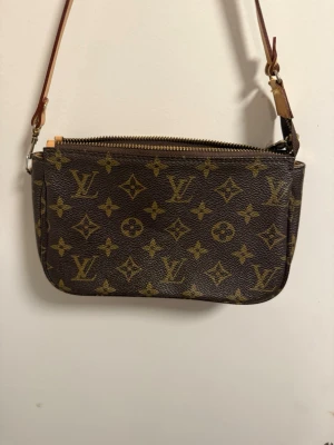 Louis Vuitton brun axelväska - Säljer en klassisk brun axelväska från Louis Vuitton med ikoniskt monogrammönster i guld. Väskan har en smidig dragkedja upptill och en justerbar rem i läder. Perfekt storlek för det viktigaste och en snygg detalj till din outfit.